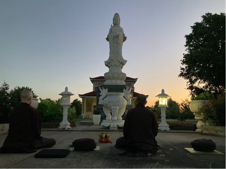 Phjoto: Two practitioners meditate outside at sunrise in front of the statue of Quan Am Bodhisattva.